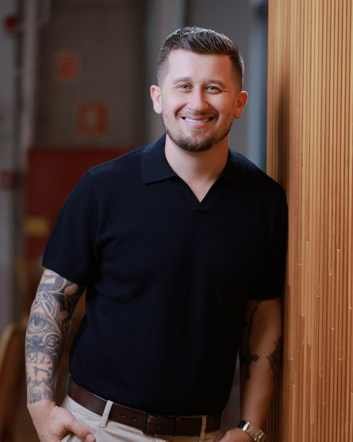 Daniel Moreira, CEO da OPEN ETHOS, sorrindo, de camisa polo escura, apoiado em um painel de madeira